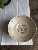 A handmade ceramic bowl with a cream-colored glaze and hand-painted brown spots, resting on a white cloth over a rustic wooden surface.