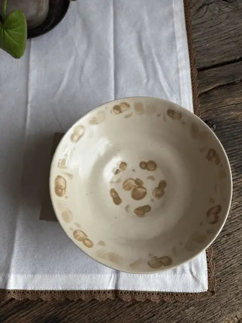 A handmade ceramic bowl with a cream-colored glaze and hand-painted brown spots, resting on a white cloth over a rustic wooden surface.