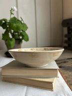 A shallow cream-colored ceramic bowl with subtle hand-painted floral patterns, resting on a stack of vintage books on a white tablecloth, with a blurred potted plant in the background.