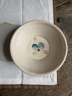 A handmade ceramic bowl in a warm cream-beige glaze, featuring a small teal and sandy painted floral motif inside the bottom. The bowl is displayed on a brown wooden trivet over a white linen cloth on a rustic wooden table.