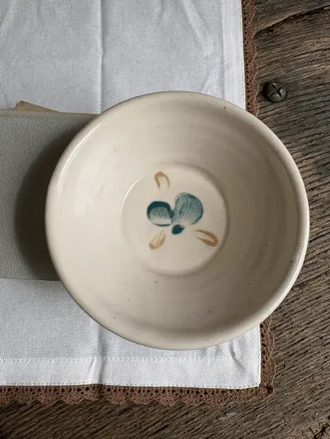 A handmade ceramic bowl in a warm cream-beige glaze, featuring a small teal and sandy painted floral motif inside the bottom. The bowl is displayed on a brown wooden trivet over a white linen cloth on a rustic wooden table.