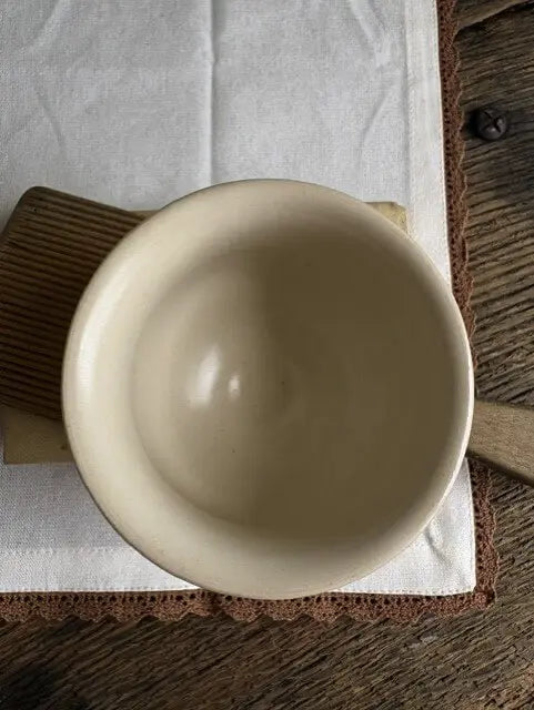 A top-down view of a light beige ceramic bowl resting on a white cloth with a brown wooden handle, placed on a rustic dark wood table.