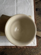 A top-down view of a light beige ceramic bowl resting on a white cloth with a brown wooden handle, placed on a rustic dark wood table.