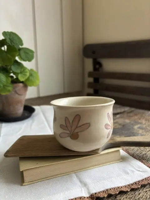 A cream-colored ceramic cup with pink floral patterns sits on top of two stacked books on a white tablecloth, with a potted plant and a wooden chair blurred in the background.