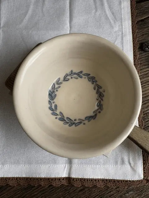 A round off-white ceramic bowl with a blue leafy wreath pattern printed inside, resting on a white cloth over a brown wooden surface.