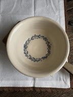A round off-white ceramic bowl with a blue leafy wreath pattern printed inside, resting on a white cloth over a brown wooden surface.