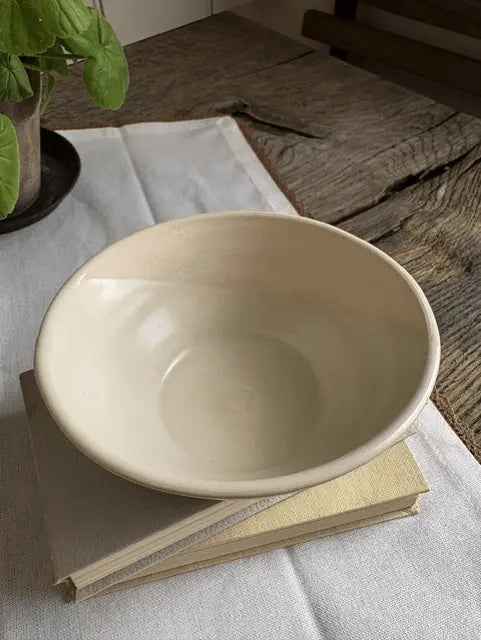 A large, shallow cream-colored ceramic bowl with a matte finish, resting on a stack of two books on a white linen cloth over a rustic wooden table.