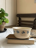 A cream-colored ceramic bowl with a blue leaf pattern, resting on a wooden book on top of another book, with a potted plant and a wooden chair in the background.