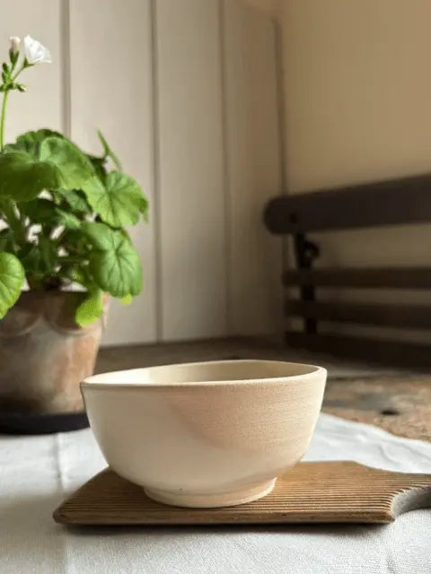 A light beige ceramic bowl with a textured matte finish, resting on a small wooden tray on a white tablecloth. A potted green plant is blurred in the background on the left, and a wooden bench is visible on the right.