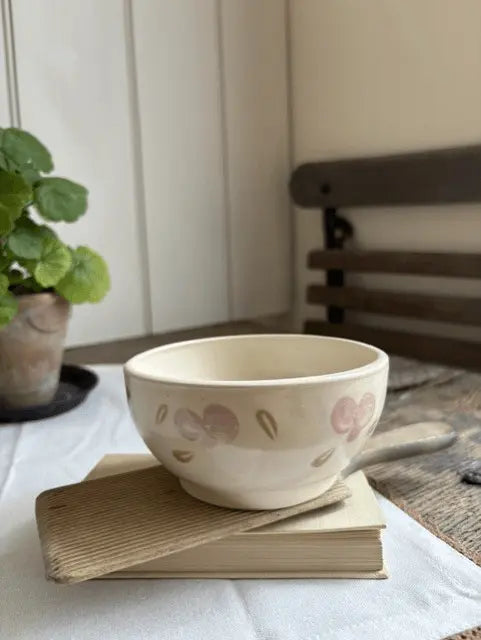 A cream-colored ceramic bowl with pink and green floral patterns, resting on a vintage book on a white tablecloth in a cozy room setting.