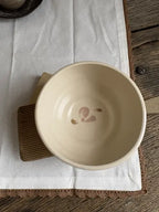 A handmade ceramic bowl in a warm cream-beige glaze, featuring a small pink painted floral motif inside the bottom. The bowl is displayed on a brown wooden trivet over a white linen cloth on a rustic wooden table.