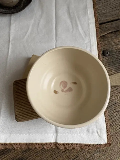 A handmade ceramic bowl in a warm cream-beige glaze, featuring a small pink painted floral motif inside the bottom. The bowl is displayed on a brown wooden trivet over a white linen cloth on a rustic wooden table.