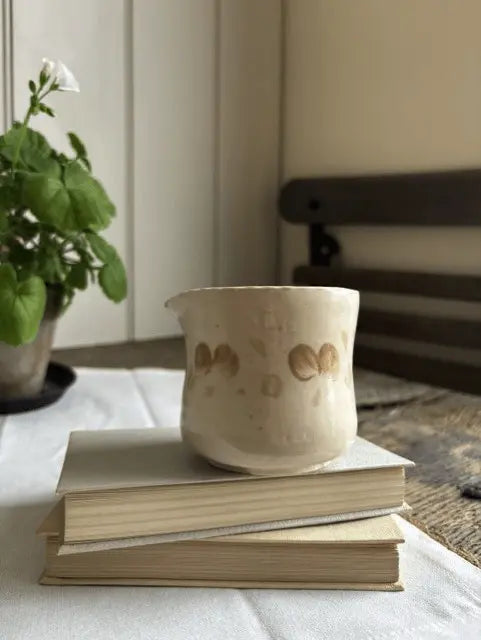A small beige ceramic cup with a glossy finish and hand-painted brown leaf motifs, resting on a stack of two vintage books on a white tablecloth. A potted green plant is visible in the soft-focus background.