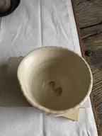 A handmade ceramic bowl with a rustic, hand-painted finish in a warm cream-beige color, resting on a white fabric runner over a wooden surface.