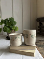 Two small handmade ceramic cups with a cream glaze and green and gold leaf patterns, displayed on a stack of books on a white tablecloth with a potted plant in the background.