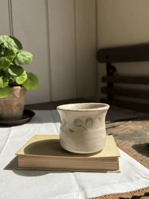 A small handmade ceramic cup with a flared rim sits on a stack of two books on a white tablecloth. The cup is off-white with hand-painted abstract motifs in muted blue, brown, and orange.