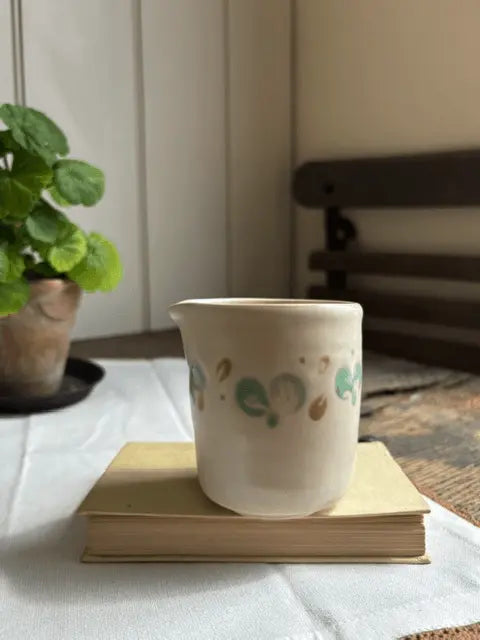 A small handmade ceramic cup with a white body and green and gold painted dots, sitting on a vintage book on a white tablecloth with a potted plant in the background.