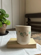 A small handmade ceramic cup with a white body and green and gold painted dots, sitting on a vintage book on a white tablecloth with a potted plant in the background.
