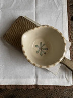 A small handmade ceramic bowl with a scalloped rim and a painted green flower motif inside, resting on a brown striped cloth over a white fabric surface.