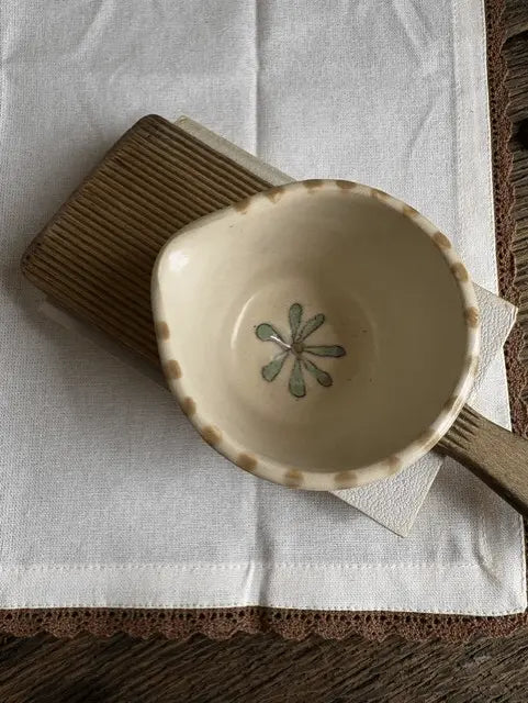 A small handmade ceramic bowl with a scalloped rim and a painted green flower motif inside, resting on a brown striped cloth over a white fabric surface.