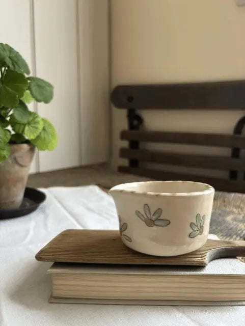 A small cream-colored ceramic cup with a hand-painted floral pattern sits on a wooden coaster resting on a book, with a potted plant and a wooden bench in the softly blurred background.
