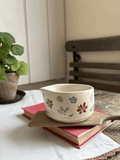 A small cream-colored ceramic cup with hand-painted floral motifs in red, blue, and green, resting on a wooden tray atop a pink book in a cozy indoor setting.