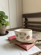 A small cream-colored ceramic cup with hand-painted floral motifs in red, blue, and green, resting on a wooden tray atop a pink book in a cozy indoor setting.