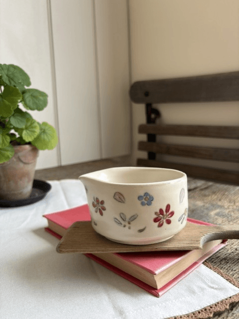A small cream-colored ceramic cup with hand-painted floral motifs in red, blue, and green, resting on a wooden tray atop a pink book in a cozy indoor setting.