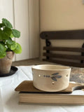 A cream-colored ceramic cup with a blue dragonfly design and small cutout details, resting on a wooden book on top of a book in a cozy indoor setting.