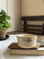 A cream-colored ceramic cup with a blue dragonfly design and small cutout details, resting on a wooden book on top of a book in a cozy indoor setting.