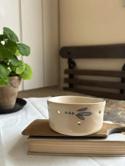 A cream-colored ceramic cup with a blue dragonfly design and small cutout details, resting on a wooden book on top of a book in a cozy indoor setting.