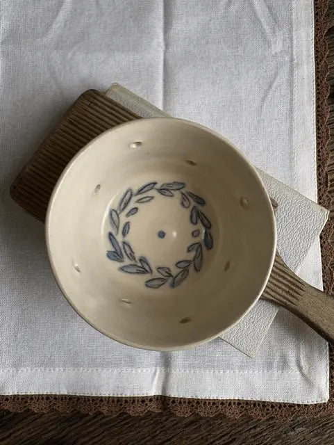 A handmade ceramic bowl with a blue leaf wreath design inside, resting on a white cloth over a brown wooden surface.