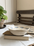 A small oval ceramic dish with a blue floral pattern sits on top of two stacked books on a white tablecloth. A potted green plant is visible on the left, and a wooden chair is blurred in the background.