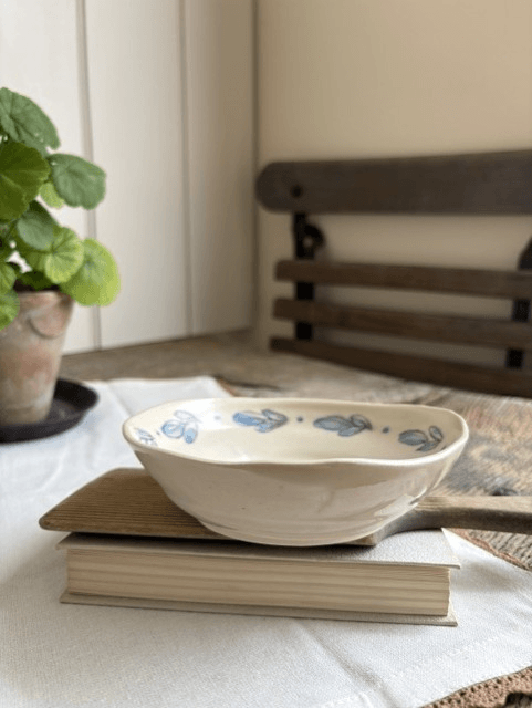 A small oval ceramic dish with a blue floral pattern sits on top of two stacked books on a white tablecloth. A potted green plant is visible on the left, and a wooden chair is blurred in the background.