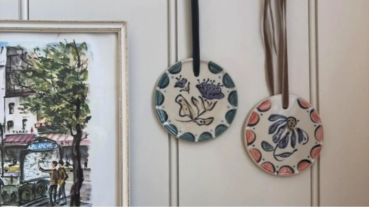 Two round ceramic decorations hang on a white panelled wall beside a framed street scene. One has a blue border and a bird with flowers; the other has a coral border and a purple flower.