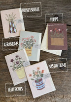 A set of botanical-themed greeting cards arranged on a rustic wooden surface, each card featuring a different flower illustration and labeled with names like Honeysuckle, Tulips, Geraniums, Nasturtiums, and Hellebores.