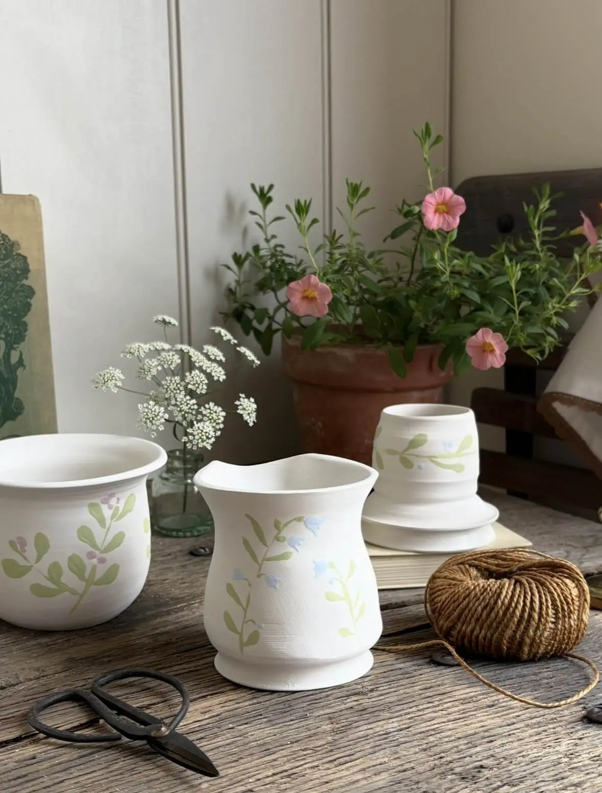 Handmade white ceramic vessels with delicate botanical designs displayed on a rustic wooden table alongside flowers and pottery tools.
