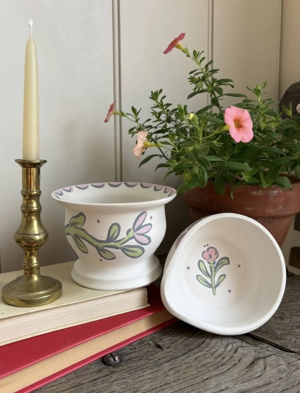 Handmade white ceramic bowl and planter with hand-painted floral designs, styled with a brass candlestick, books and flowers on a rustic wooden table.