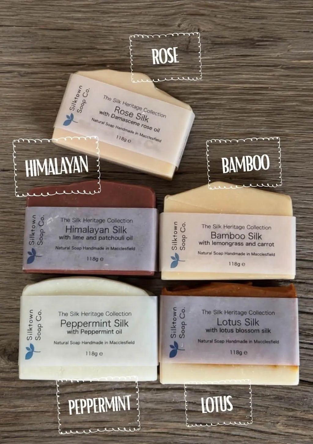 Six bars of silk soap arranged on a dark wood background, each labeled with its scent name: ROSE, HIMALAYAN, BAMBOO, PEPPERMINT, LOTUS, and one bar with no visible text label. The soap bars are shown in different colors including cream, pinkish-beige, brown, and white, with white paper labels.