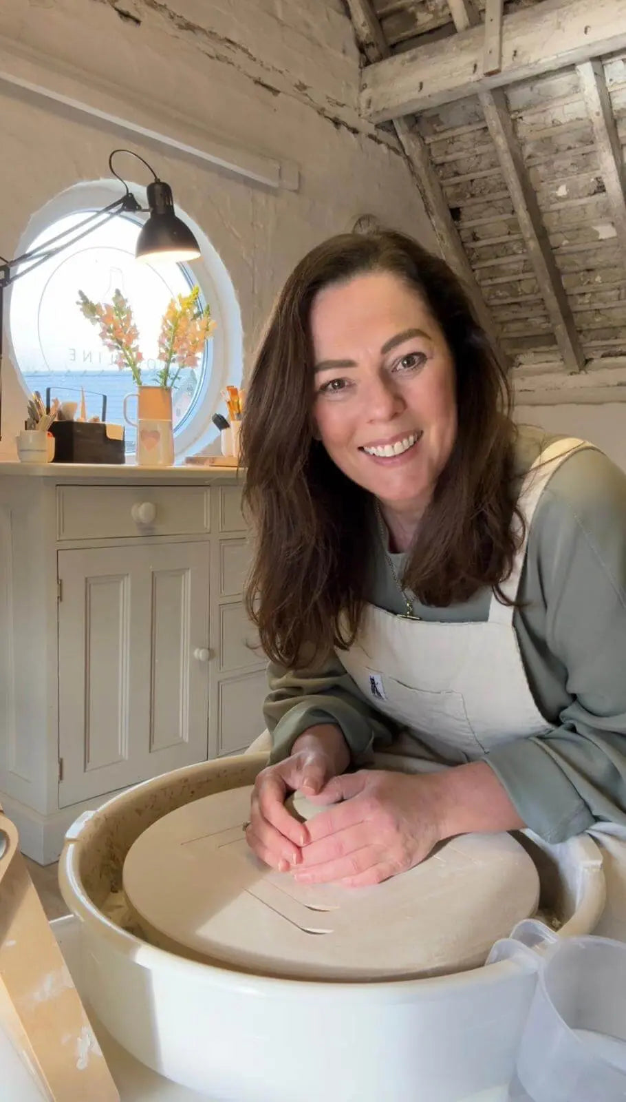 A smiling Zoe sits at a pottery wheel, working with clay inside a rustic, sunlit studio with exposed wooden ceiling beams and white walls.